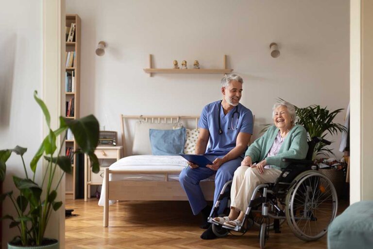 An elderly woman holds hands with a younger visitor in a comfortable assisted living room.